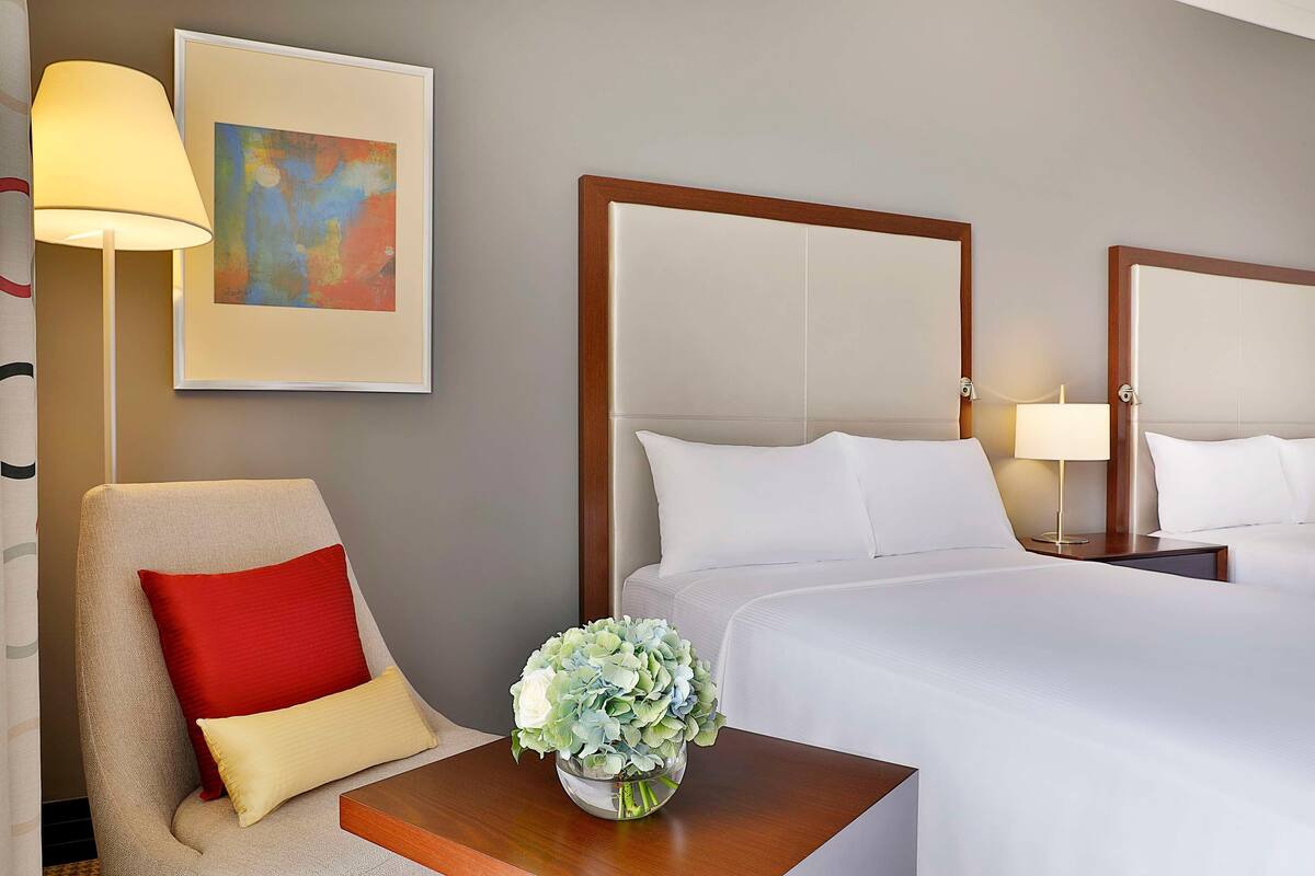 A hotel room with a bed, a chair with a red pillow, a bedside table with a vase of flowers, a lamp, and a framed abstract painting on the wall.