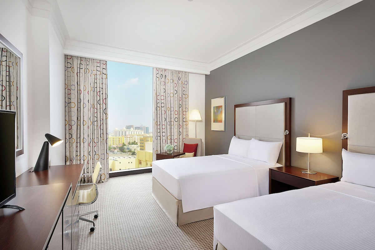 A hotel room with two beds, a desk, a chair, a TV, and a view of the city.