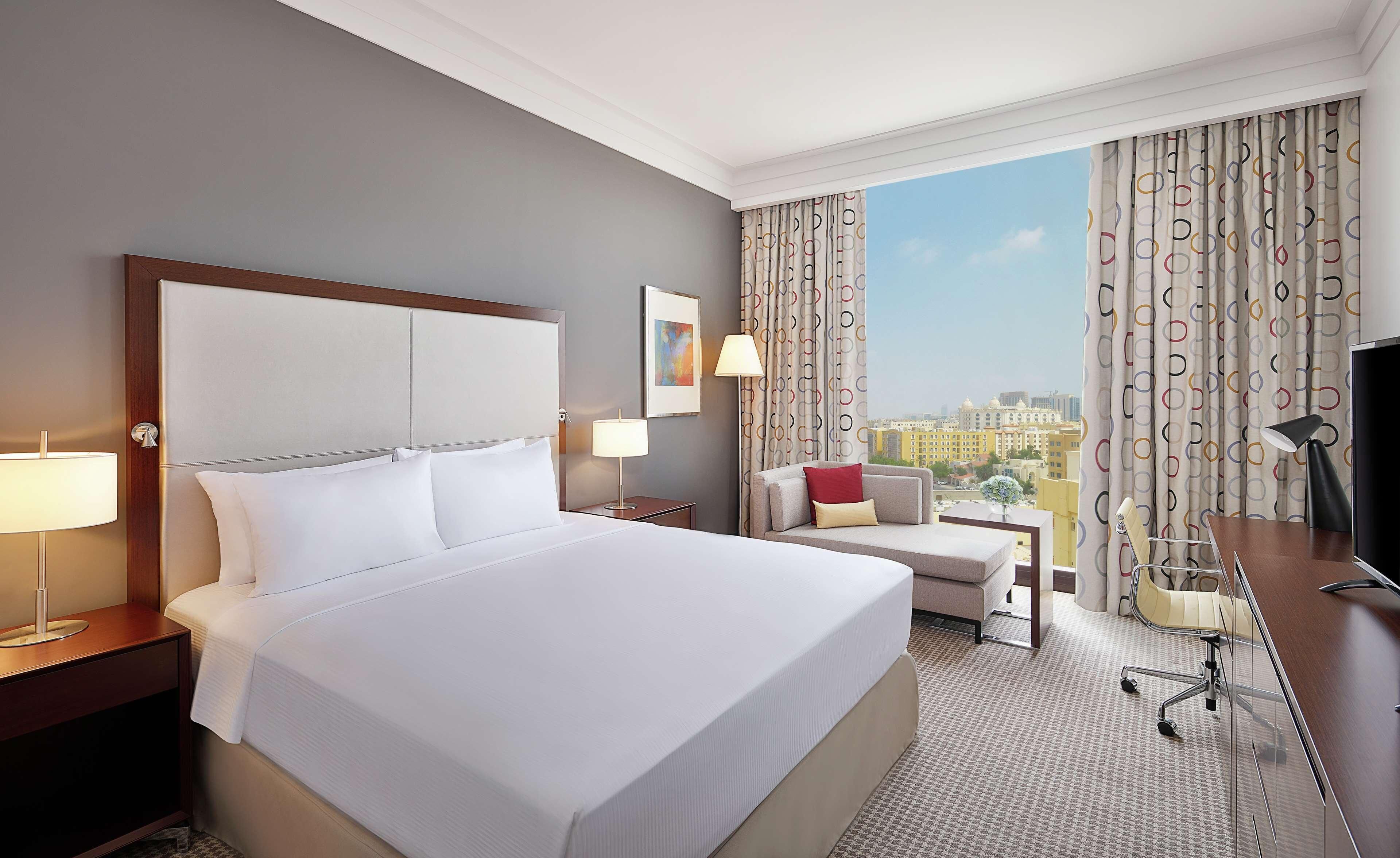 A hotel room with a large bed, a desk, a chair, a lamp, and a view of the city.