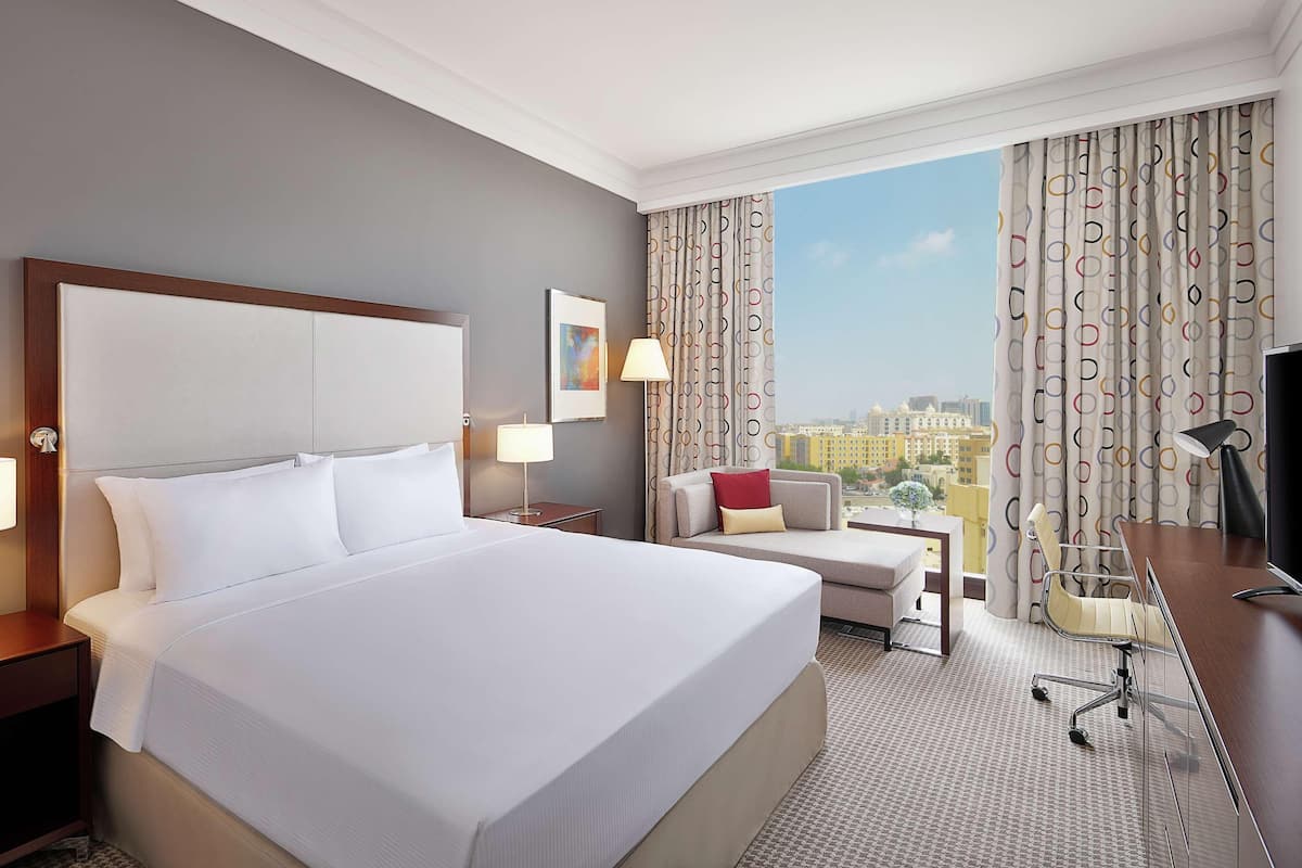 A hotel room with a large bed, a desk, a chair, a lamp, and a view of the city.