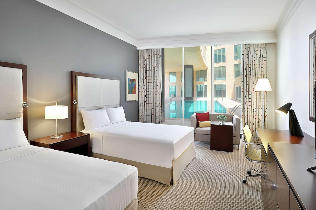 A hotel room with two beds, a desk, a chair, a TV, and a view of a swimming pool.