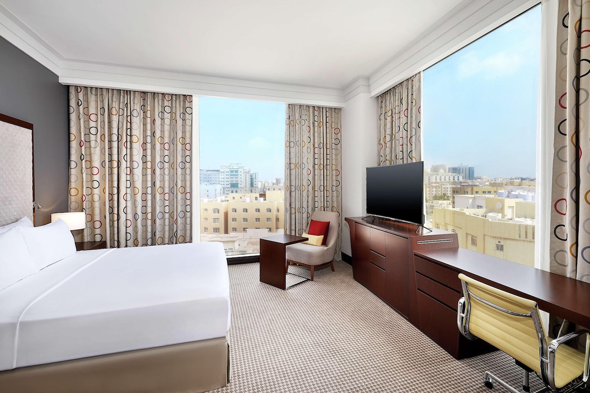A hotel room with a large bed, a desk, a chair, a TV, and a view of the city.