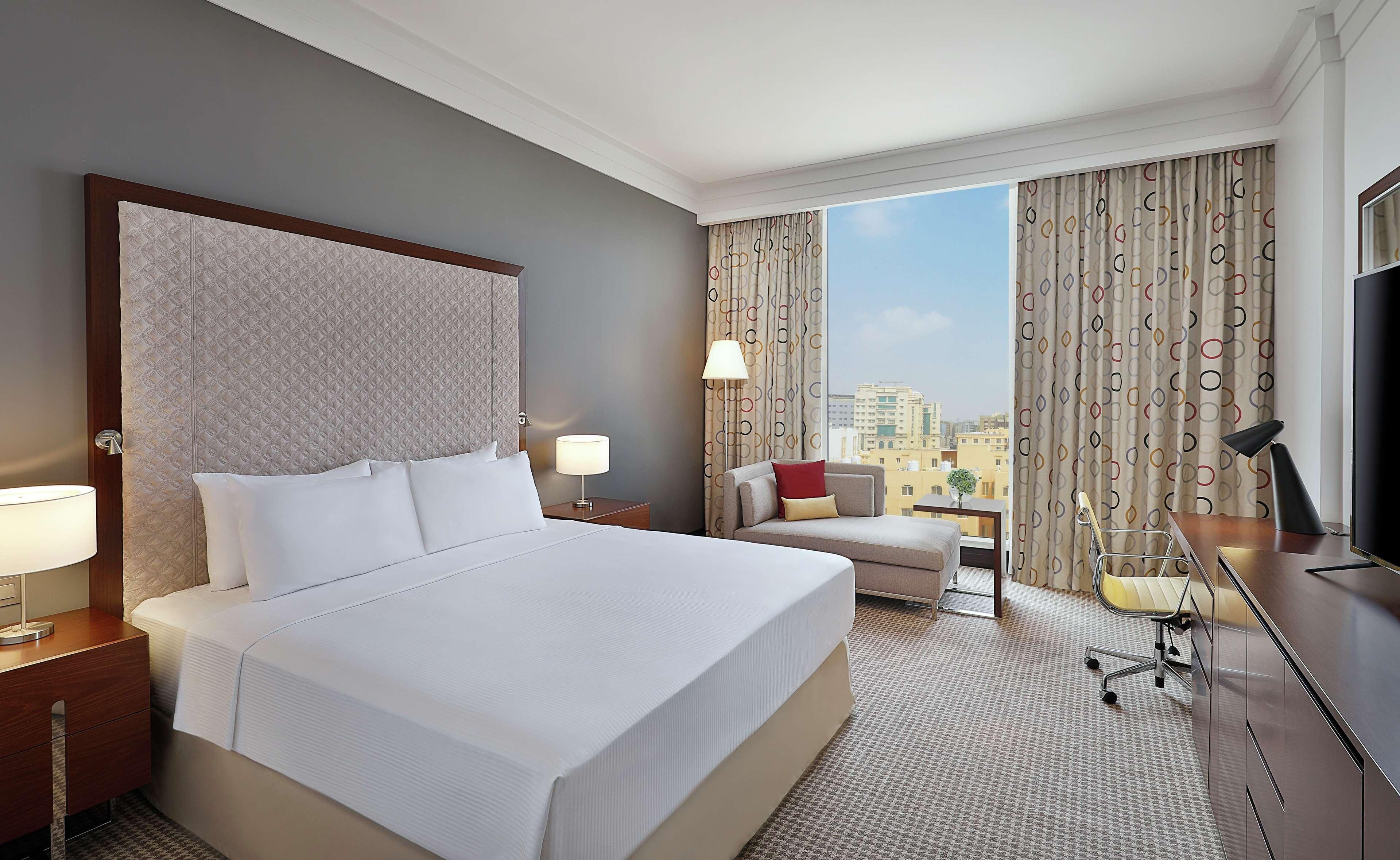 A hotel room with a large bed, a desk with a lamp, a chair, and a TV.