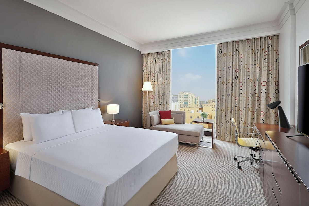 A hotel room with a large bed, a desk with a lamp, a chair, and a TV.