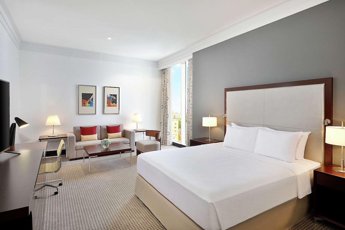 A hotel room with a large bed, a desk, a chair, two bedside lamps, a small table with a lamp, and two framed pictures on the wall.