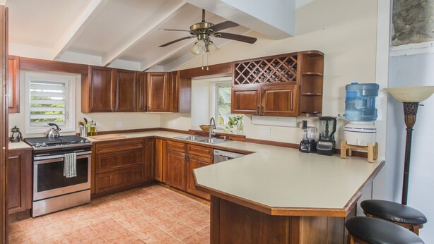 A kitchen with wooden cabinets, a stove, a dishwasher, a water dispenser, and a ceiling fan. - Feather Leaf Inn (Frederiksted)