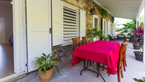 A covered outdoor dining area with a red table and chairs, potted plants, and a stone wall.