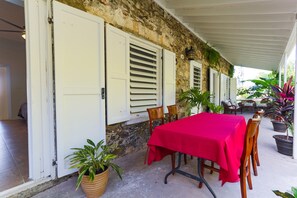 A covered outdoor dining area with a red table and chairs, potted plants, and a stone wall.