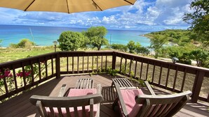 A wooden deck with two chairs and a large umbrella, overlooking a coastal view with trees and a clear sky.