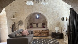 Um quarto aconchegante com parede de pedra, cama com cabeceira de madeira, sofá cinza, tapete estampado e uma pequena mesa com abajur.