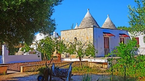 Un bâtiment en pierre, doté d’une façade blanche et d’un toit recouvert de tuiles rouges, entouré de verdure et agrémenté d’une brouette.