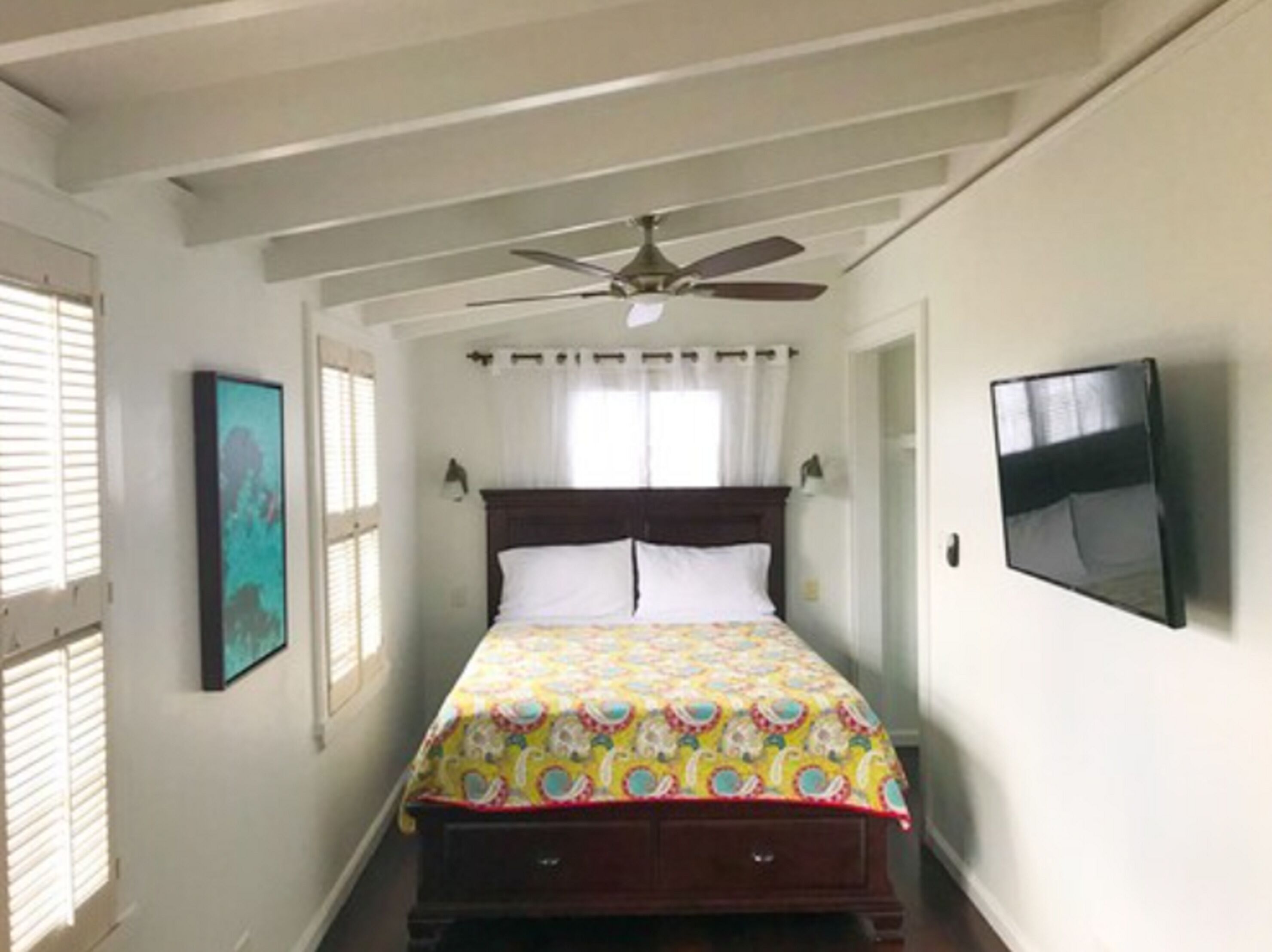 Une chambre à coucher avec un lit, un ventilateur de plafond, un téléviseur fixé au mur et une fenêtre avec des stores.