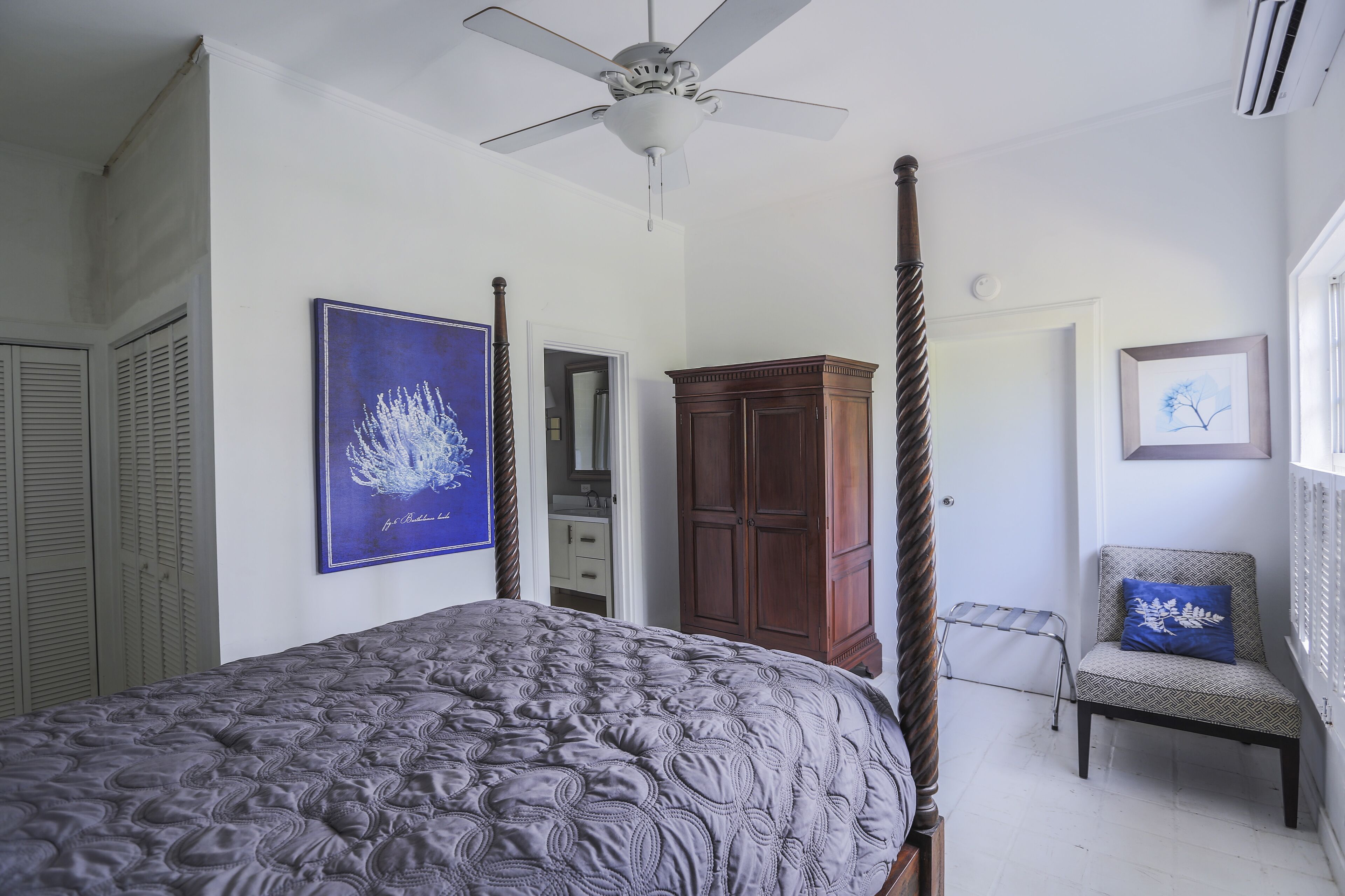 A bedroom with a four-poster bed, a ceiling fan, a wardrobe, a chair, and a framed picture on the wall.