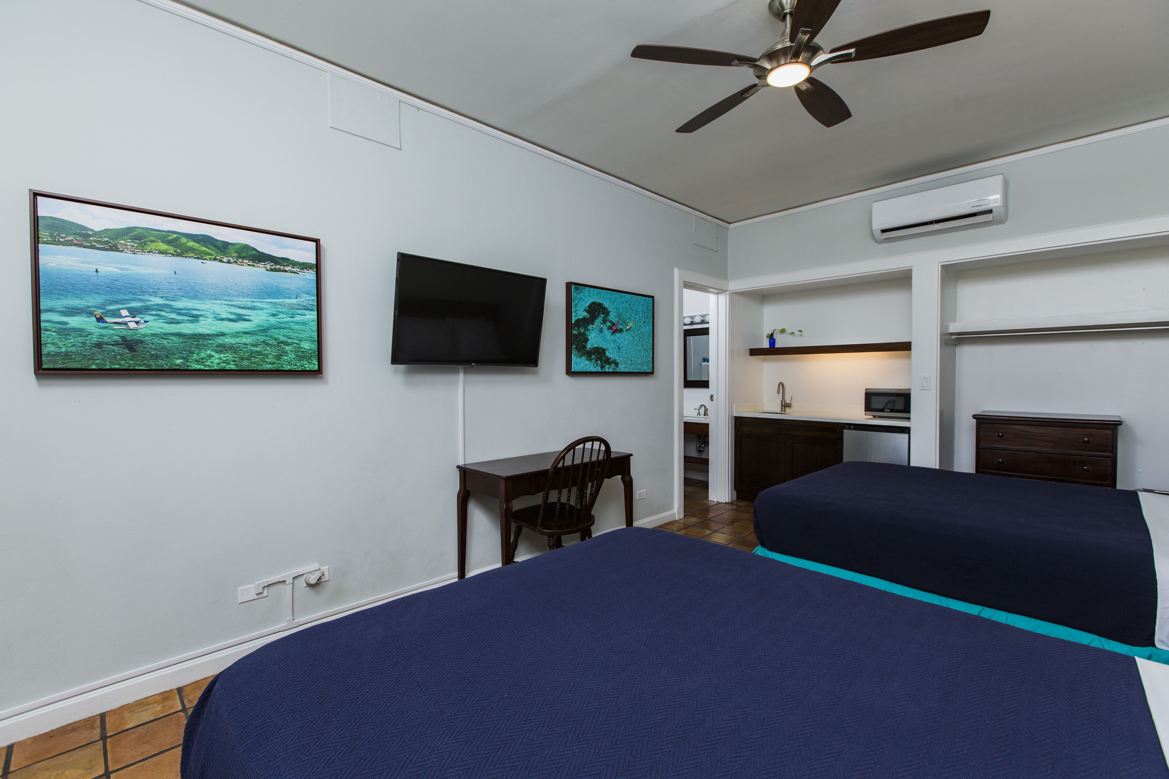 Quarto de hotel com duas camas, uma escrivaninha, uma cadeira, uma televisão e um ventilador de teto.