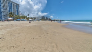 A wide sandy beach with palm trees and beach umbrellas, a large hotel building, and a clear blue sky. - Beautiful Isla Verde Apartment by the Beach! (Carolina)