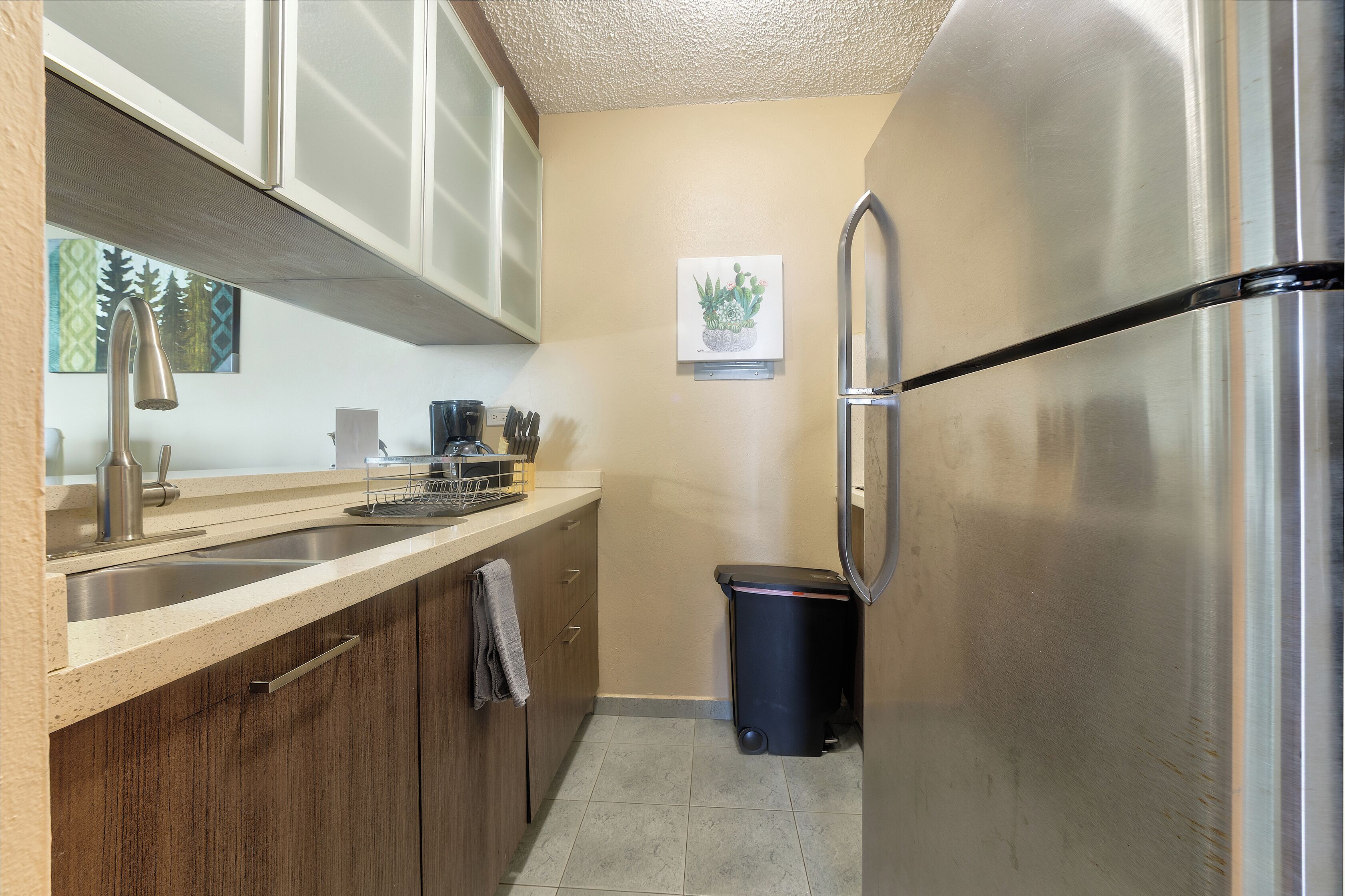 A kitchen with a stainless steel refrigerator, a sink, wooden cabinets, and a trash bin.