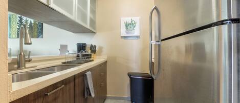 A kitchen with a stainless steel refrigerator, a sink, wooden cabinets, and a trash bin.