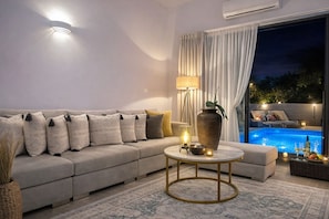 A modern living room with a beige sofa, a round coffee table, and a large vase. There is a pool visible through the window. - Villa Almihauss Adults-Only  (Afantou)