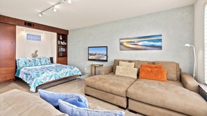 A modern living room with a sofa, a bed with a blue floral bedspread, a bookshelf, and a flat-screen TV mounted on the wall.