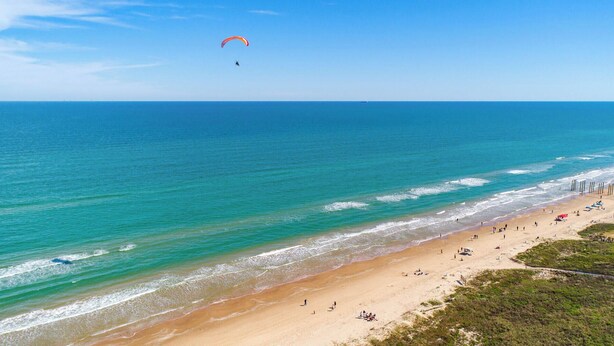A beach with a clear view of the ocean, a person paragliding, and a sandy shore with people and umbrellas.