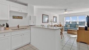 A modern kitchen with white cabinetry, a central island, and a dining area with wicker chairs. There is a large window providing natural light and a view of the city.