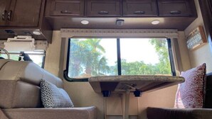 A compact RV interior with a kitchenette, a dining area, and a window providing a view of the outdoors.