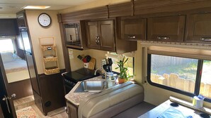 A compact RV kitchen with a microwave, sink, and stove, featuring a window with a view of a wooden fence.