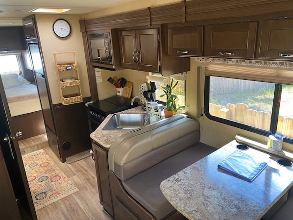 A compact RV kitchen with a microwave, sink, and stove, featuring a window with a view of a wooden fence.