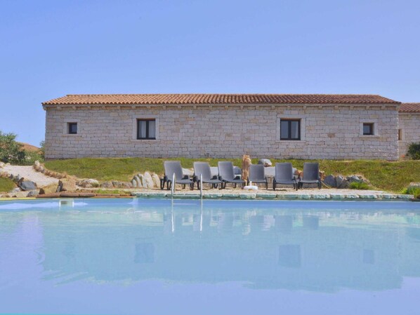 A stone building with a tiled roof, a pool, and several lounge chairs.
