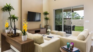 A modern living room with a flat-screen TV, comfortable seating, a coffee table with books and decorative items, and a view of the outdoors through large sliding glass doors. - Awesome 4 Bedroom Villa type home near Disney (Kissimmee)