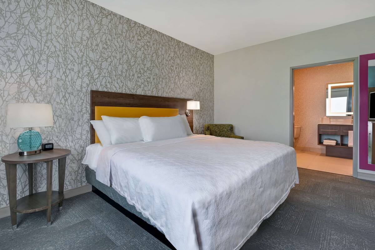 A hotel room with a large bed, a bedside table with a lamp, and a bathroom visible through an open door.