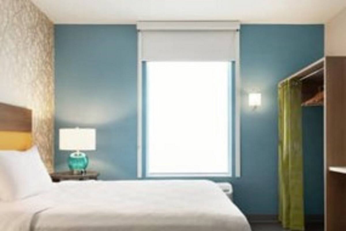 A hotel room with a bed, a nightstand with a lamp, a window with blinds, and a closet with a green curtain.