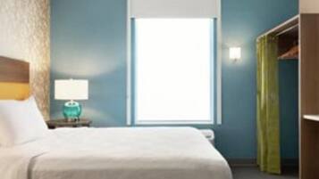 A hotel room with a bed, a nightstand with a lamp, a window with blinds, and a closet with a green curtain.