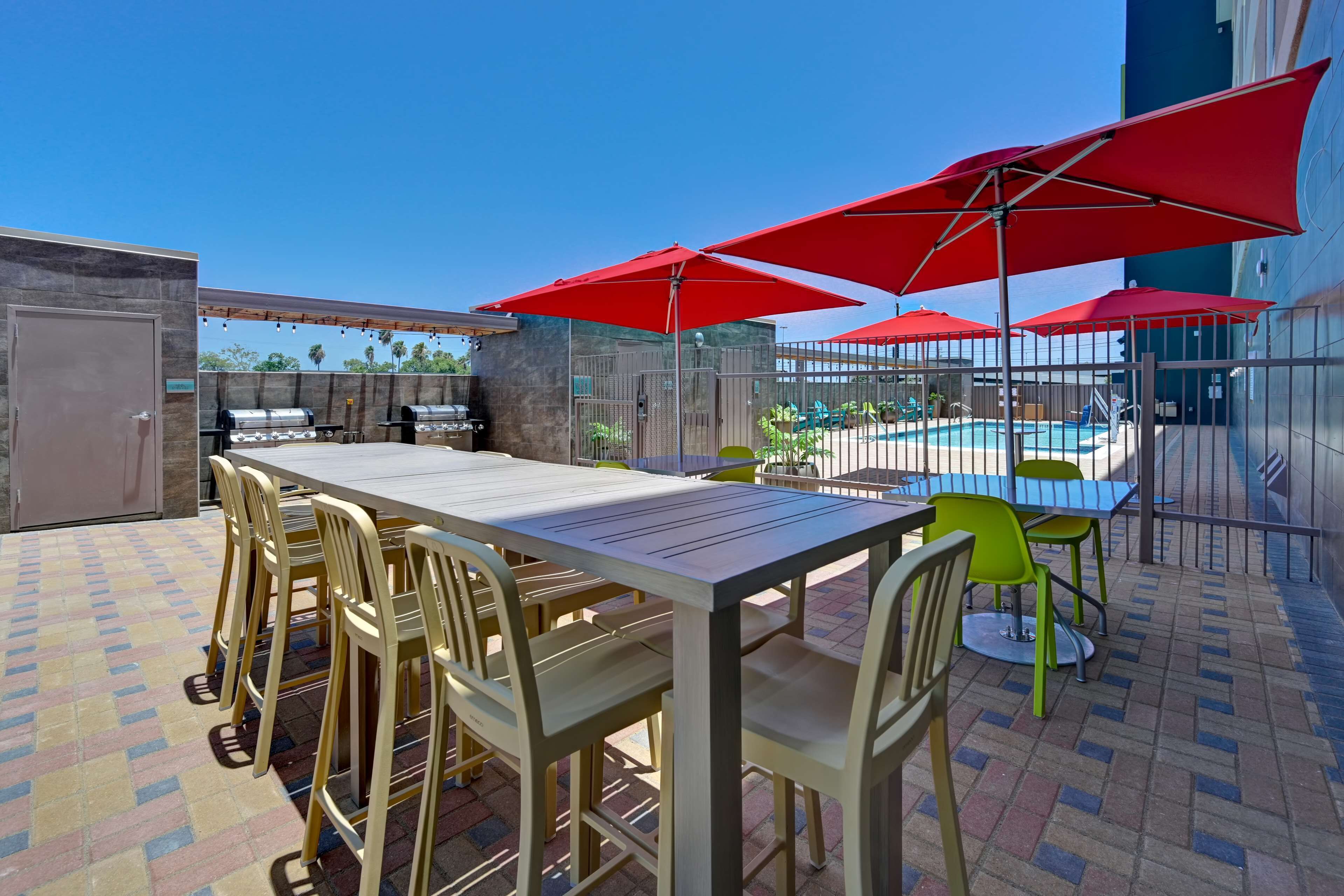 An outdoor patio with a long table and chairs, red umbrellas, and a pool in the background.