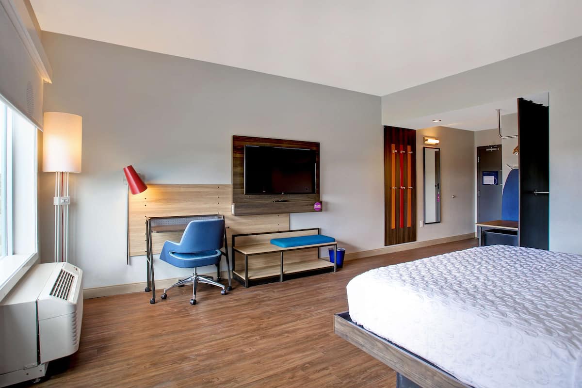 A hotel room with a bed, a desk with a chair, a TV mounted on the wall, and a window.