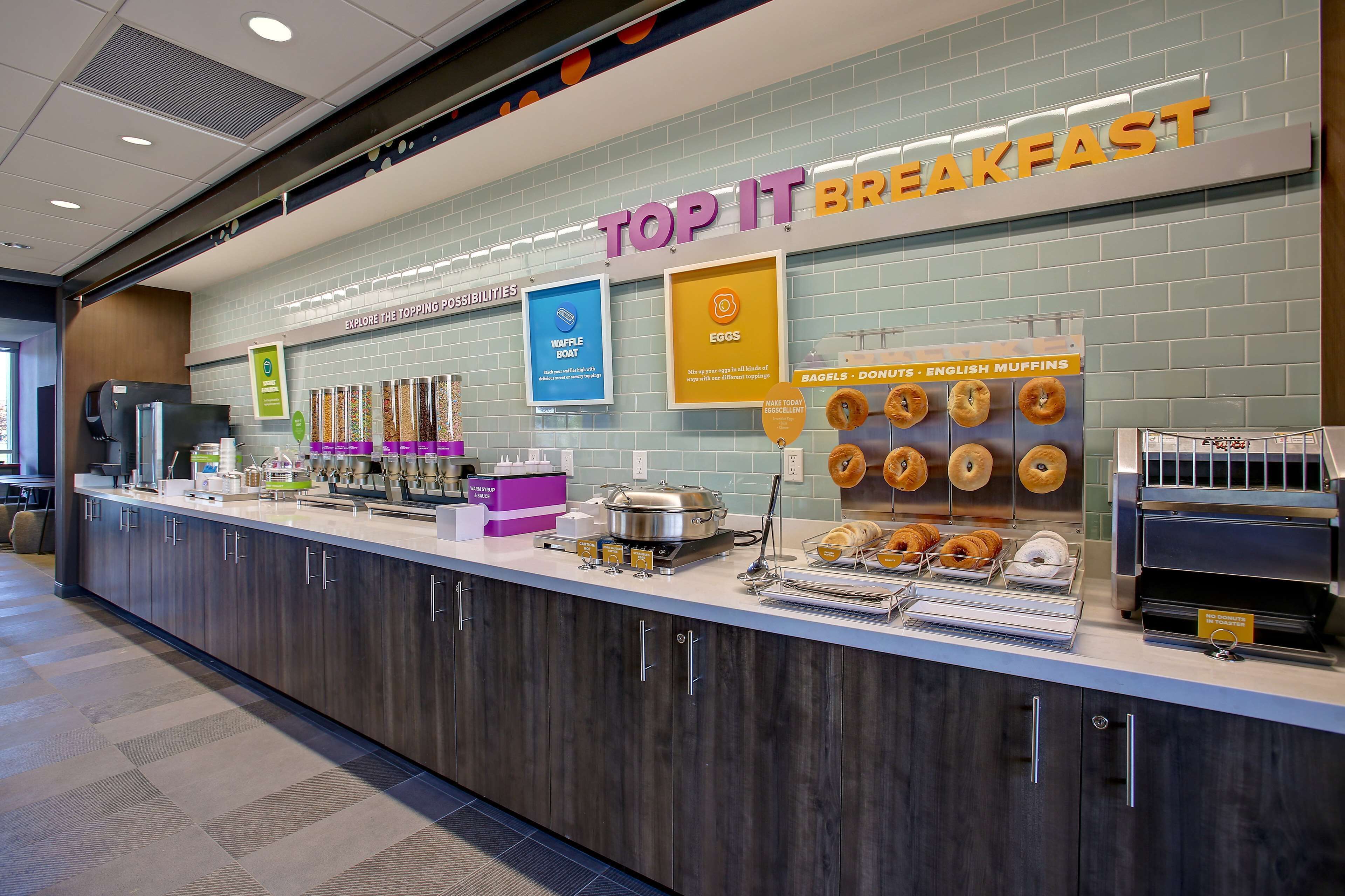 A breakfast counter with a sign reading "TOP IT BREAKFAST" and various food options like waffles, eggs, and donuts.