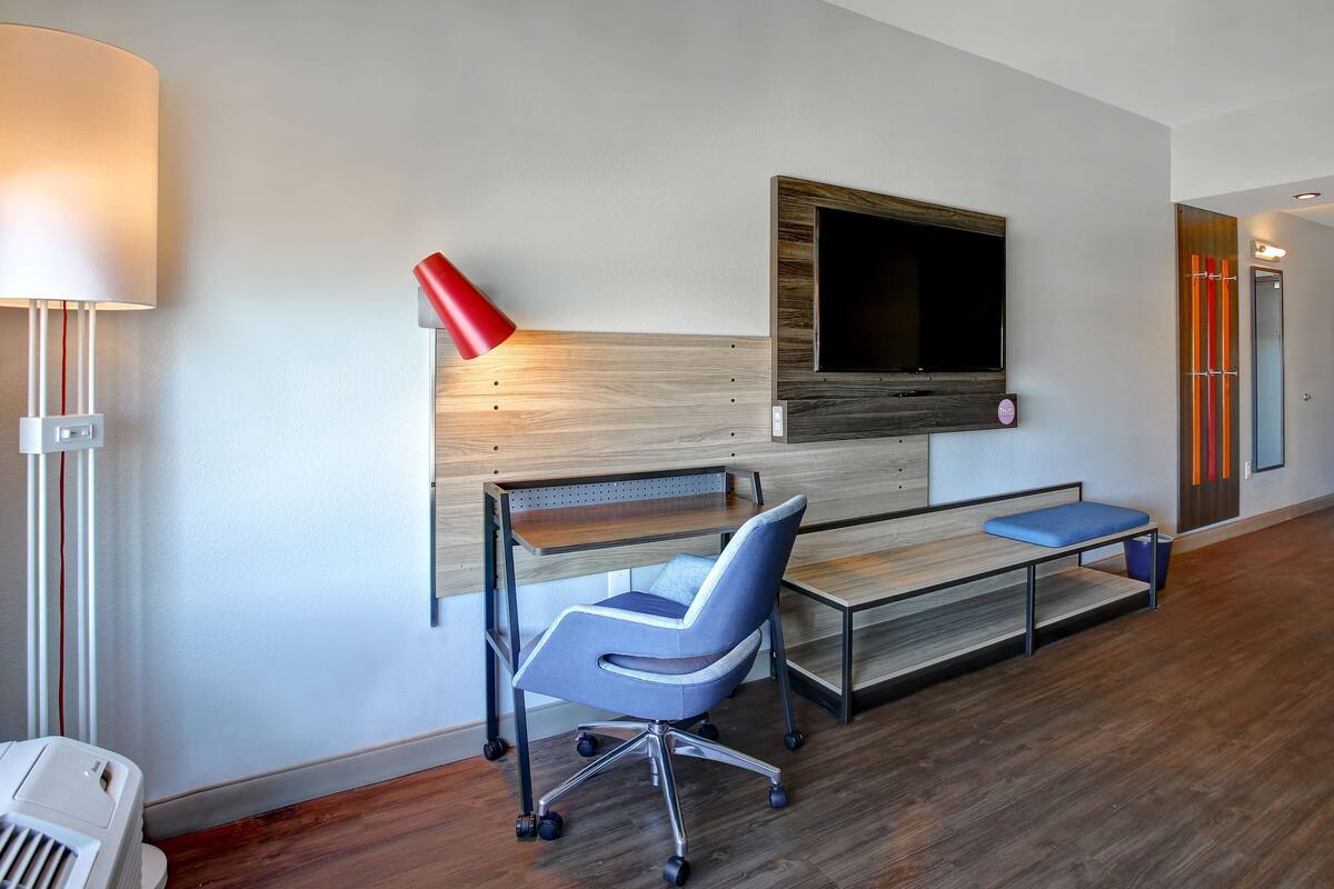 A modern hotel room with a wooden desk, a blue office chair, a TV mounted on the wall, and a red lamp.