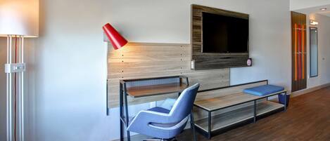 Habitación de hotel moderna con un escritorio de madera, una silla de oficina azul, un televisor montado en la pared y una lámpara roja.
