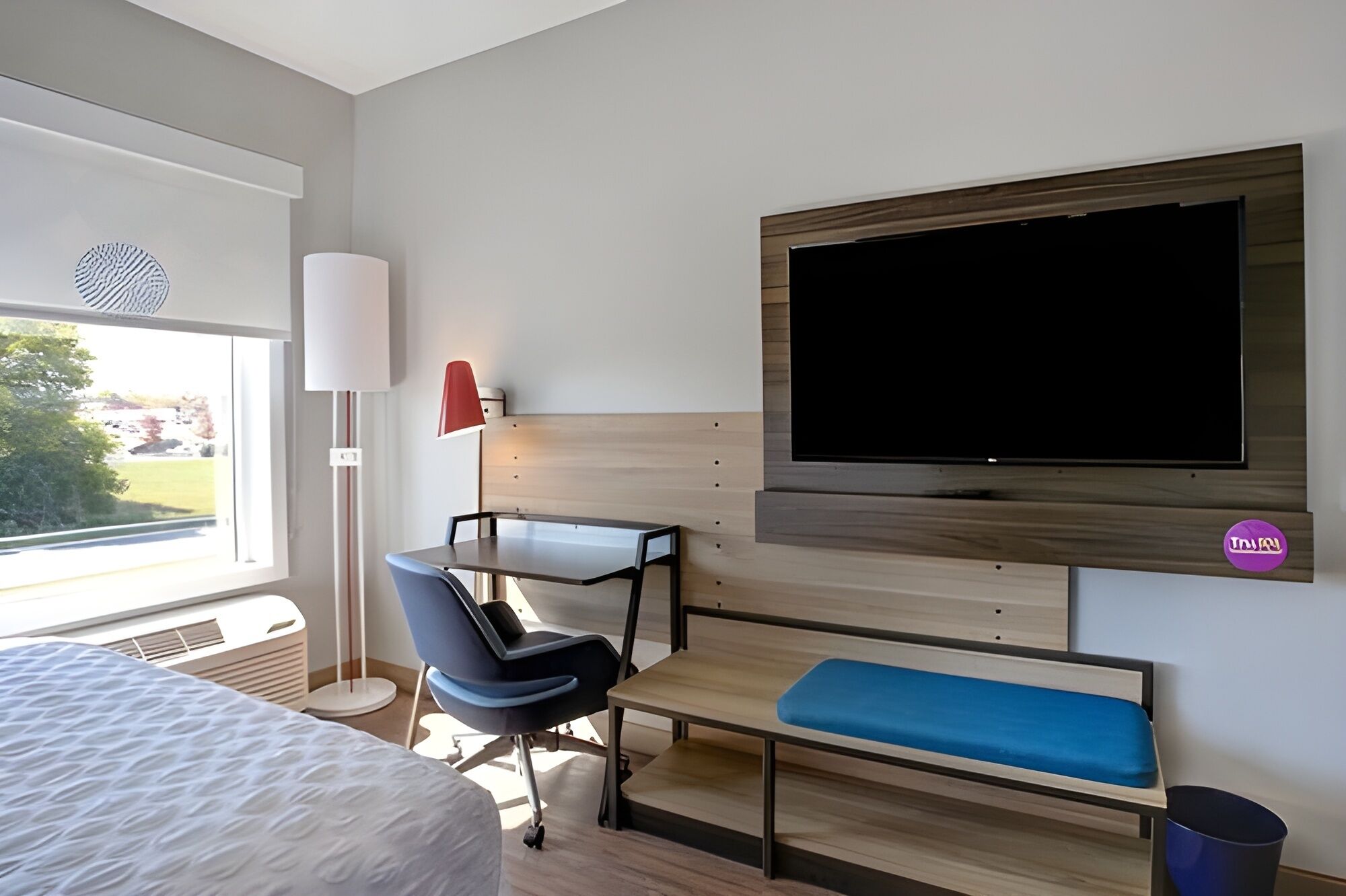 Ett hotellrum med en säng, ett skrivbord med en stol, en tv, en bänk och ett fönster.