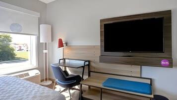 Ett hotellrum med en säng, ett skrivbord med en stol, en tv, en bänk och ett fönster.