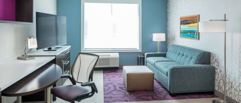 A hotel room with a desk, chair, sofa, and a large window.