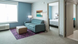 Ein Hotelzimmer mit blauer Wand, einem türkisblauen Sofa, einem kleinen Hocker, einem lila gemusterten Teppich, einem Gemälde, einem Bett und einer Tür zu einem anderen Raum.