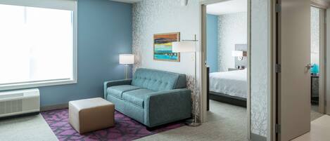 A hotel room with a blue wall, a teal sofa, a small ottoman, a purple patterned rug, a painting, a bed, and a door leading to another room.