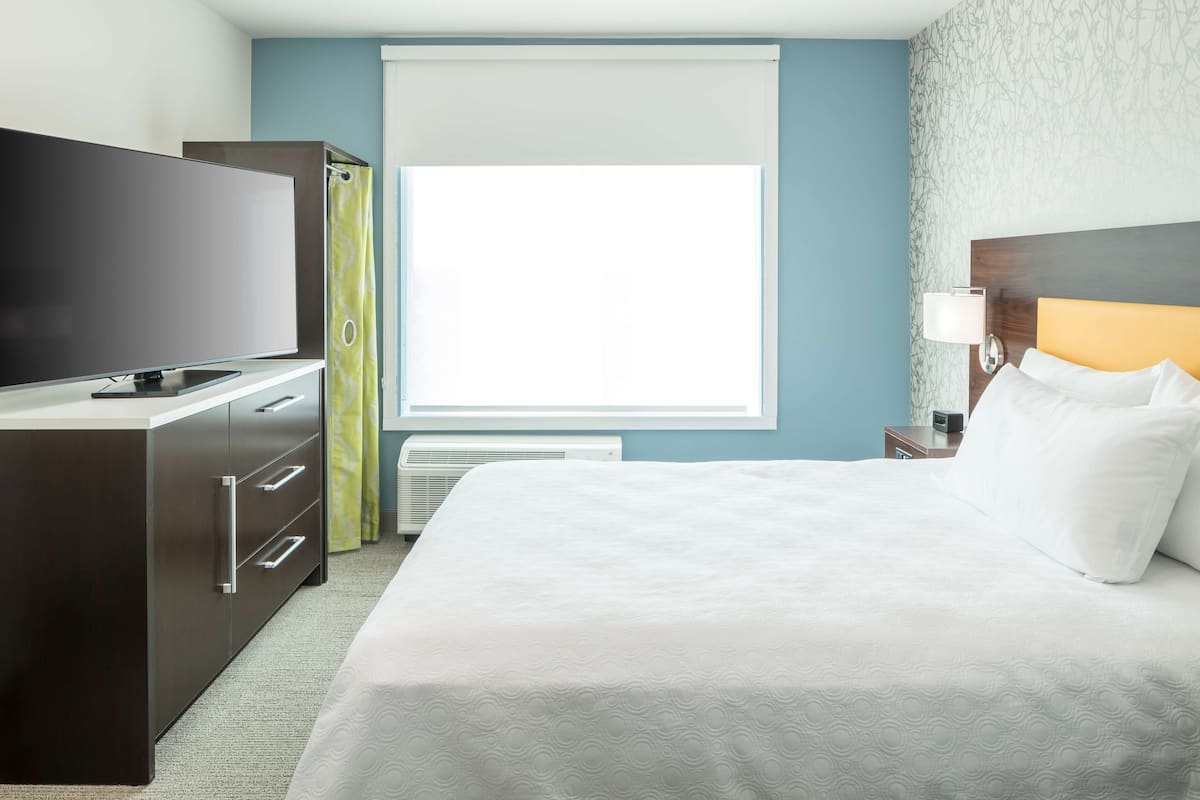 Ein Hotelzimmer mit einem Bett, einem Fernseher, einem Kleiderschrank, einem Fenster mit Vorhängen und einem Nachttisch mit Lampe.