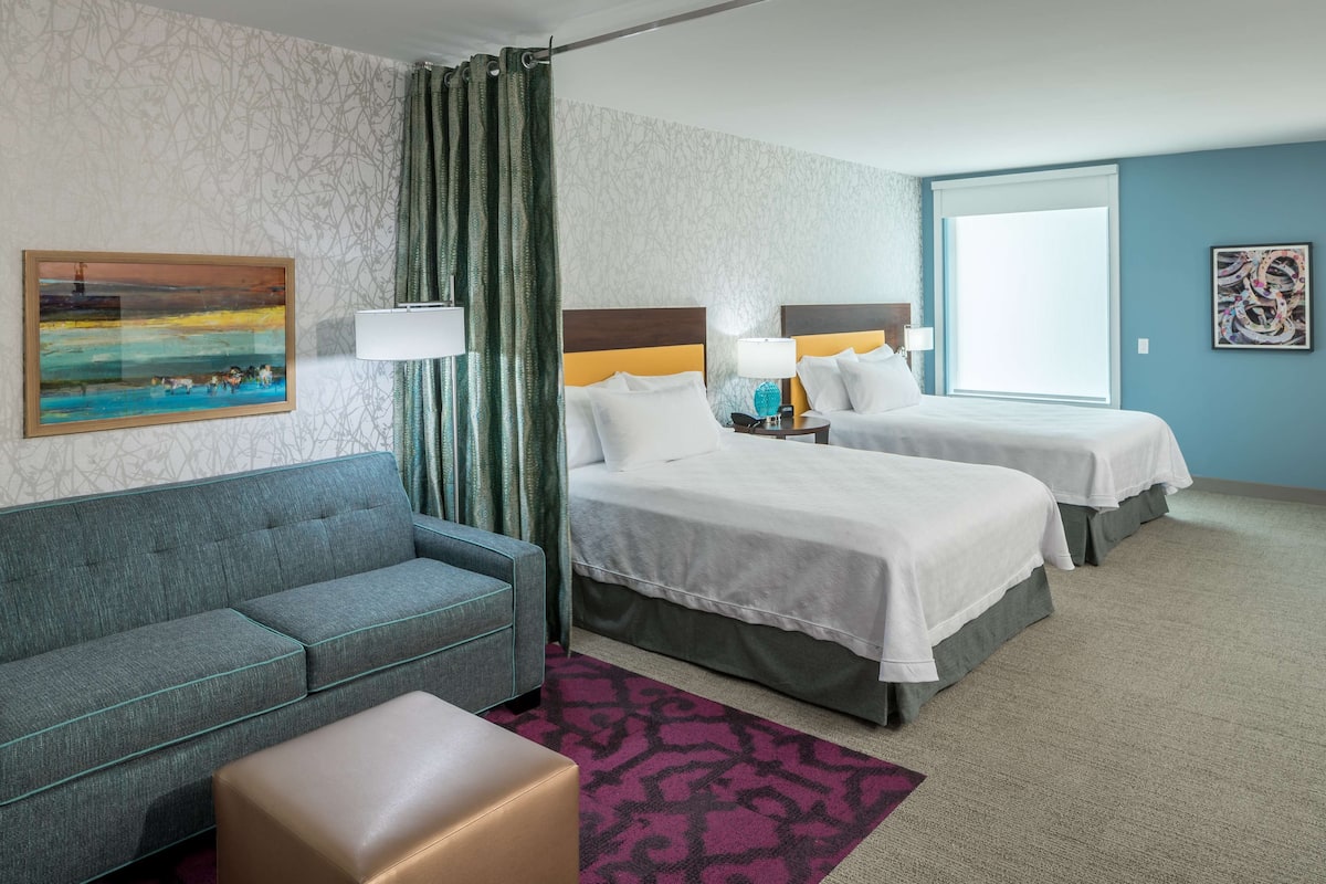 Ein Hotelzimmer mit zwei Betten, einem Sofa, einem kleinen Tisch und einem Fenster mit Vorhängen.