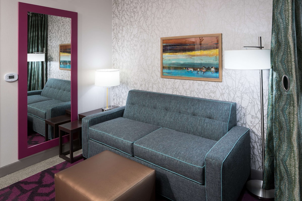Ein Hotelzimmer mit grauem Sofa, einem kleinen Tisch, einem Spiegel, einem Gemälde, einer Lampe und einem gemusterten Teppich.