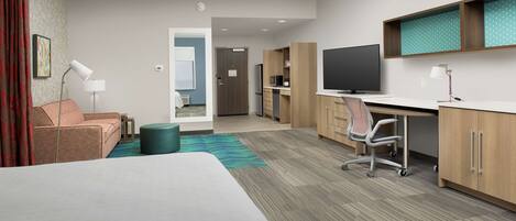 A hotel room with a bed, a desk, a chair, a TV, and a kitchenette.