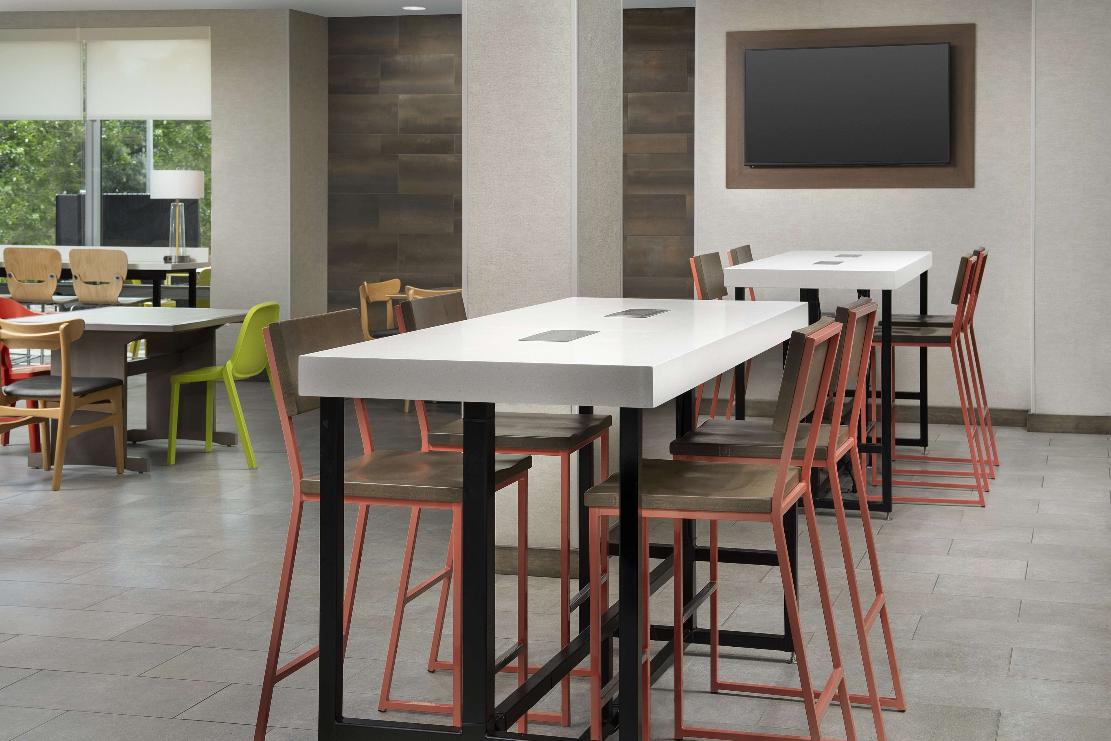 A modern conference room with white tables and red chairs, large windows, and a flat-screen TV on the wall.