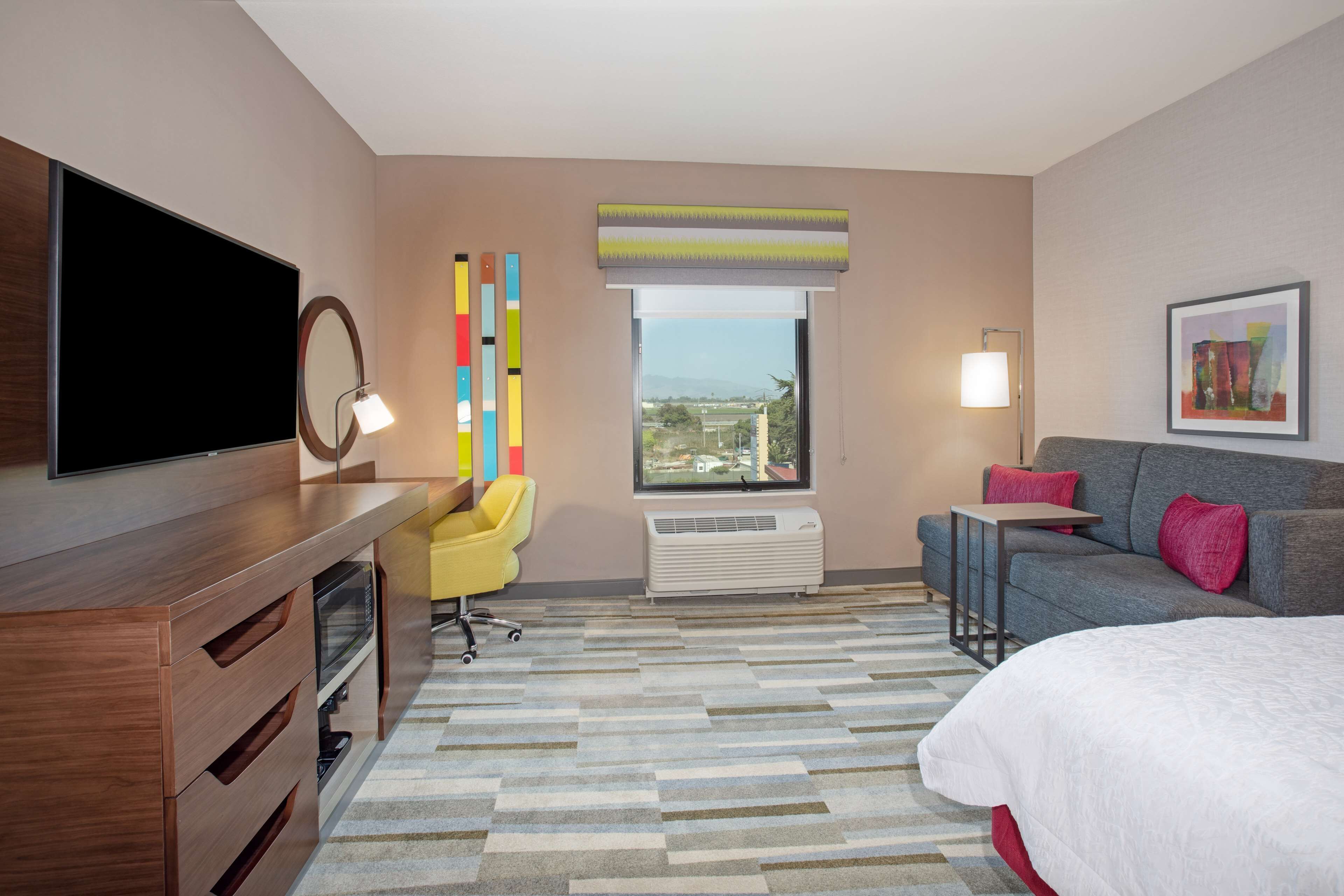 Habitación de hotel con cama, escritorio con televisor de pantalla plana, silla, sofá, ventana con vistas y aire acondicionado instalado en la pared.