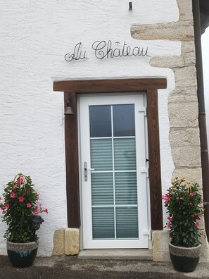 A white door with a wooden frame and a glass panel, flanked by two potted plants with pink flowers, and the text "Au Chateau" above the door. - Duplex with a terrace and a carport (Lajoux)